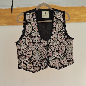 Puede incluir: Un chaleco negro con intrincados bordados multicolores y pequeñas lentejuelas en un patrón repetitivo de flores y cachemira. La prenda tiene un escote en V, un borde de pompones negros y una etiqueta rectangular que dice Persian Harmony.