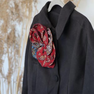 Könnte beinhalten: Schwarze Wolljacke mit einer dekorativen roten Stoffblume mit Muster, die an das Revers genäht ist. Die Blume hat blaue, goldene und weiße Akzente. Die Jacke ist geknöpft und hängt an einem grauen Kleiderbügel.