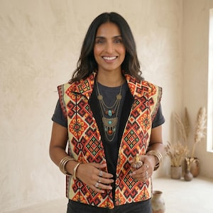 Könnte beinhalten: Eine gemusterte Weste mit einem geometrischen Design in Rot, Orange und Gelb. Die Weste wird über einem dunkelgrauen T-Shirt getragen und mit goldenen Armbändern und Ringen kombiniert. Eine mehrreihige Halskette mit türkisfarbenen Akzenten vervollständigt den Look.