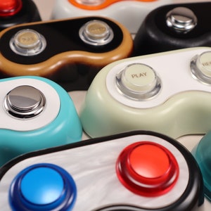 May include: A collection of retro-style audio devices in various colors, including black, teal, and white. Each device has two buttons, one labeled "PLAY" and the other "RECORD", with a shiny silver button on top. The devices have a rounded, ergonomic design.