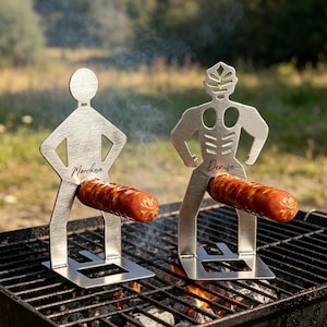 Könnte beinhalten: Zwei silberfarbene Metallspieße in Form menschlicher Figuren auf einem schwarzen Holzkohlegrill. Jede Figur hält eine braune Wurst. Die linke Figur ist mit dem Namen Marchesa und die rechte Figur mit dem Namen Denise graviert.