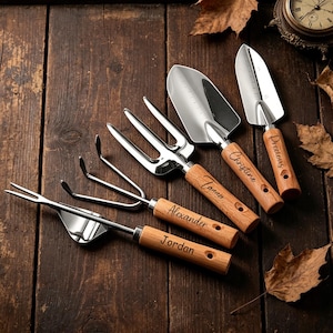 Könnte beinhalten: Ein Set aus fünf Gartenwerkzeugen aus Edelstahl mit hellen Holzgriffen, die jeweils mit einem Namen graviert sind: Jordan, Alexander, Tanner, Christine und Precious. Das Set enthält einen Unkrautstecher, einen Rechen, eine Gabel und zwei Schaufeln.