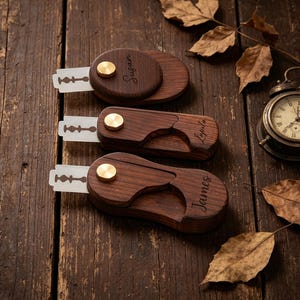 Könnte beinhalten: Drei personalisierte Rasiererhalter aus Holz mit freiliegenden Klingen, jeweils mit einem messingfarbenen Knopf. Die Halter sind mit den Namen Susan, Lyla und James graviert. Sie liegen auf einer rustikalen Holzoberfläche, mit einer Vintage-Uhr und getrockneten Blättern.