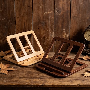 May include: Two wooden book stands, one light-colored and one dark brown, are displayed on a wooden surface. Each stand has a sloped design with a ledge at the bottom. A vintage clock and autumn leaves are in the background.