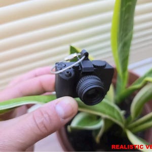 May include: A small, black camera with a lens and a silver wire loop is held in a hand. The camera is positioned in front of a green and yellow plant. The text "REALISTIC VERSION" is at the bottom of the image.