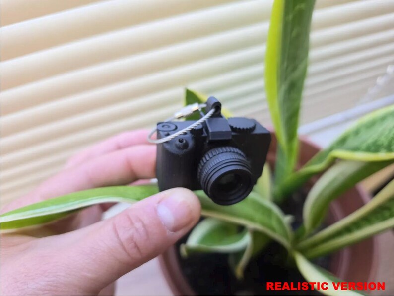 May include: A small, black camera with a lens and a silver wire loop is held in a hand. The camera is positioned in front of a green and yellow plant. The text "REALISTIC VERSION" is at the bottom of the image.
