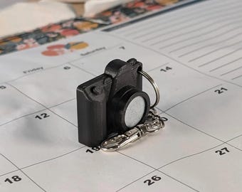 May include: A black camera-shaped keyring with a silver key ring and clasp. The keyring is on a calendar page with the text "SLIM VERSION" in red. A white ceramic item is in the background.
