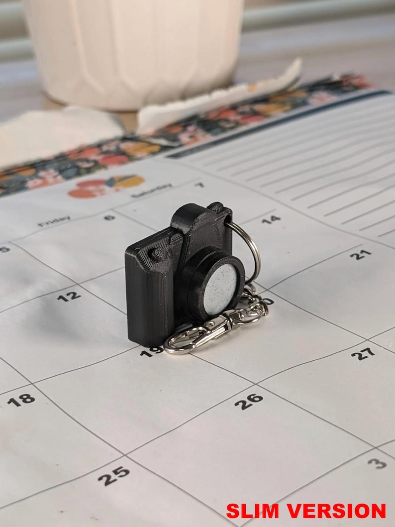 May include: A black camera-shaped keychain with a silver key ring and clasp. The keychain is on a calendar page with the text "SLIM VERSION" in red. A white ceramic item is in the background.