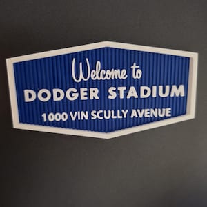 Puede incluir: Un letrero azul y blanco que dice "Welcome to Dodger Stadium 1000 Vin Scully Avenue." El letrero tiene un fondo azul texturizado y un borde blanco. El texto es blanco, con un "Welcome to" en cursiva y el resto en letras mayúsculas.