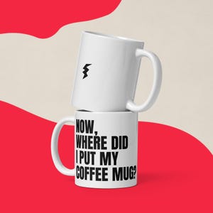 May include: Two white ceramic coffee mugs stacked on top of each other. The top mug has a black lightning bolt graphic. The bottom mug has the text: "NOW, WHERE DID I PUT MY COFFEE MUG?" The background is red and beige.