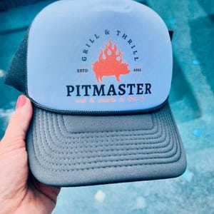 May include: A light blue and gray trucker hat with a logo that says "Grill & Thrill" and "Pitmaster" with a pig in flames. The text below the logo says "rub it, smoke it, love it".
