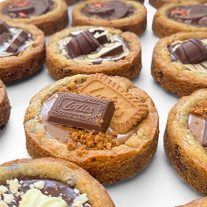 May include: A selection of mini cookie tarts, each with a different topping, are arranged on a white surface. The toppings include chocolate bars, biscuits, and other sweet treats. The tarts have a golden-brown crust, creating a tempting display of baked goods.