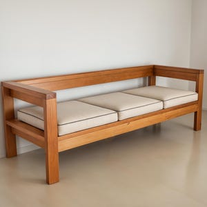 Puede incluir: Un sofá de madera de tres plazas con un acabado en madera natural y cojines blancos. El sofá tiene un diseño simple y moderno con un marco rectangular y líneas rectas.