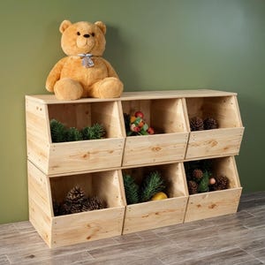 May include: A light-colored wooden storage unit with six open compartments, each filled with decorative items like pinecones and greenery. A plush teddy bear sits on top. The unit is made of natural wood.