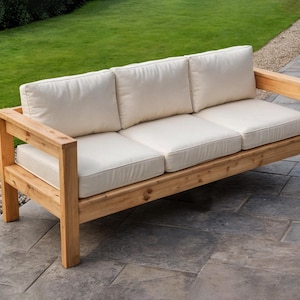 Könnte beinhalten: Ein dreisitziges Outdoor-Sofa mit einem Rahmen aus Naturholz und cremefarbenen Kissen. Das Sofa hat ein rechteckiges Design mit Armlehnen und steht auf einer Steinterrasse. Im Hintergrund sind grünes Gras und einige kleine Steine zu sehen. Ca. 200 cm lang.