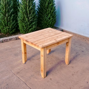 May include: A small, wooden table with a natural finish. The table has a square top and four legs. It is sitting on a concrete patio.
