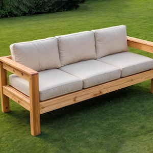 May include: A three-seater outdoor sofa with a light wood frame and beige cushions. The sofa has a rectangular design with armrests and is placed on a green lawn. The cushions are thick and appear comfortable.
