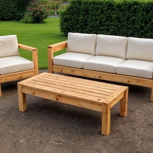 Könnte beinhalten: Ein Gartenmöbel-Set aus Holz mit einem Dreisitzer-Sofa, einem Sessel und einem rechteckigen Couchtisch. Die Rahmen sind aus hellem Holz gefertigt, und die Kissen sind cremefarben. Das Set steht auf einer Terrasse mit Rasen und Büschen im Hintergrund.