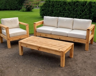 May include: A wooden outdoor furniture set featuring a three-seat sofa, an armchair, and a rectangular coffee table. The frames are made of light-colored wood, and the cushions are off-white. The set is placed on a patio with a green lawn and bushes in the background.