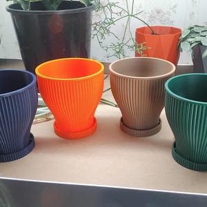 May include: A selection of ribbed plant pots in shades of navy blue, orange, beige, and green, each with a matching saucer. The pots are of varying sizes. Other potted plants are in the background.