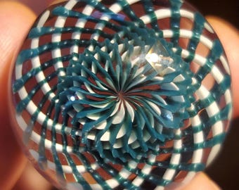 May include: A clear glass marble with an intricate design. The marble features a teal and white spiral pattern at the center, surrounded by a grid of teal, white, and brown lines. The design creates a mesmerizing optical effect.