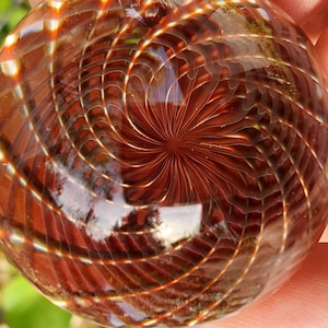 May include: A clear, spherical glass object with an intricate, spiraling design in shades of brown and amber. The pattern resembles a web or a series of interconnected lines, creating a captivating visual effect. The glass reflects light, enhancing the depth and complexity of the design.