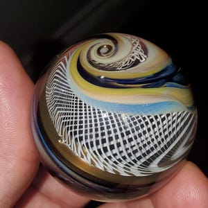 May include: A glass marble with a swirling pattern of blue, yellow, white, and gold. The marble has a grid-like pattern on the bottom half.