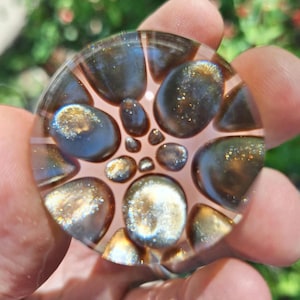 May include: A clear, round glass object with a pattern of dark brown and gold-flecked shapes. The shapes are arranged in a spiral pattern on a light pink background. The glass has a reflective, shiny surface.