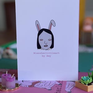 May include: A white greeting card featuring a centered illustration of a person with black hair wearing pink and white rabbit ears. Below the illustration, the text reads Blessherlittleart by Amy in a simple, dark, sans-serif font.