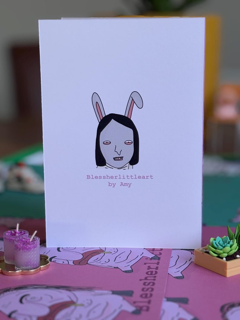 May include: A white greeting card featuring a centered illustration of a person with black hair wearing pink and white rabbit ears. Below the illustration, the text reads Blessherlittleart by Amy in a simple, dark, sans-serif font.
