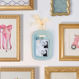 Könnte beinhalten: Ein hellblauer Bilderrahmen mit Griff und gelbem Band, der ein Schwarz-Weiß-Foto enthält. Der Rahmen ist von anderen gerahmten Kunstwerken umgeben, darunter ein rosa High-Heel-Gemälde und eine rosa Roller-Illustration.