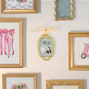 Könnte beinhalten: Eine Sammlung gerahmter Kunstwerke und ein dekorativer Fotorahmen. Der Fotorahmen ist oval, hellblau und goldfarben umrandet, mit einem cremefarbenen Band. Die Kunstwerke umfassen ein Gemälde mit rosa High Heels, eine Roller-Illustration und Blumendrucke, alle in goldenen Rahmen.