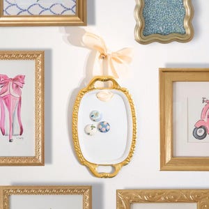 May include: A gold-framed, oval-shaped white magnetic board with three decorative magnets. The board is suspended by a peach-colored ribbon and bow. Surrounding the board are several gold-framed pictures with various images.