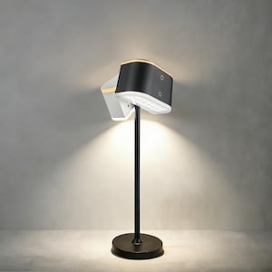 May include: A modern black and white table lamp with a unique design. The lamp features a black base and stem, with a dual-light head. The head has a black exterior and a white interior, with a warm light emanating from it. The lamp is approximately 24 inches tall.
