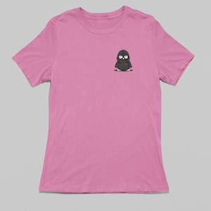 Puede incluir: Una camiseta rosa con un pequeño pingüino de dibujos animados negro que lleva una sudadera con capucha negra. El pingüino tiene ojos blancos y un pequeño pico negro.