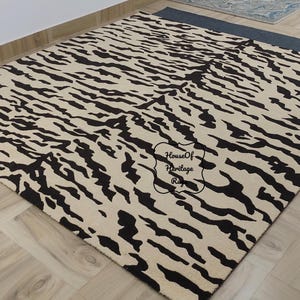 May include: A rectangular area rug featuring a cream-coloured base with a repeating black tiger stripe pattern. The rug displays a central black stamp with the text House Of Heritage Rugs. The design covers the entire surface of the floor covering.