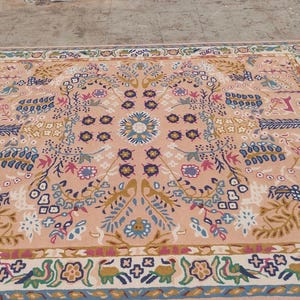 Peut inclure: Tapis rectangulaire avec un motif floral dans les tons p&ecirc;che, bleu, vert et marron. Le tapis pr&eacute;sente un m&eacute;daillon central et une bordure avec des motifs r&eacute;p&eacute;titifs. Le tapis est probablement en laine ou dans une mati&egrave;re similaire.