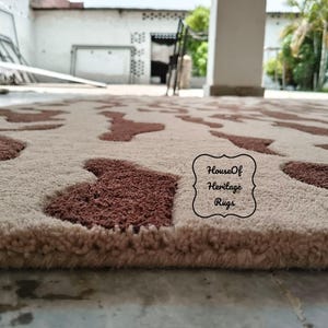 May include: A rectangular area rug featuring a cream-colored base with a repeating pattern of dark brown, irregular, cow-hide style patches. The rug has a thick, tufted pile texture and includes a black logo stamp reading House Of Heritage Rugs.