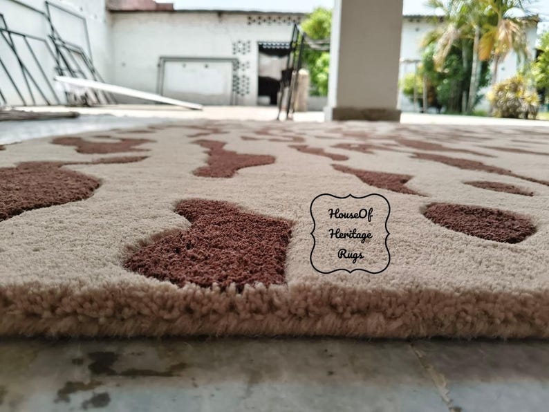 May include: A rectangular area rug featuring a cream-colored base with a repeating pattern of dark brown, irregular, cow-hide style patches. The rug has a thick, tufted pile texture and includes a black logo stamp reading House Of Heritage Rugs.