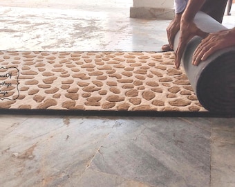May include: A long runner rug with a beige and brown pebble-like pattern. The rug features a black border and a logo that reads House Of Heritage Rugs. The rug is partially rolled up, showing a dark gray underside and a textured surface.