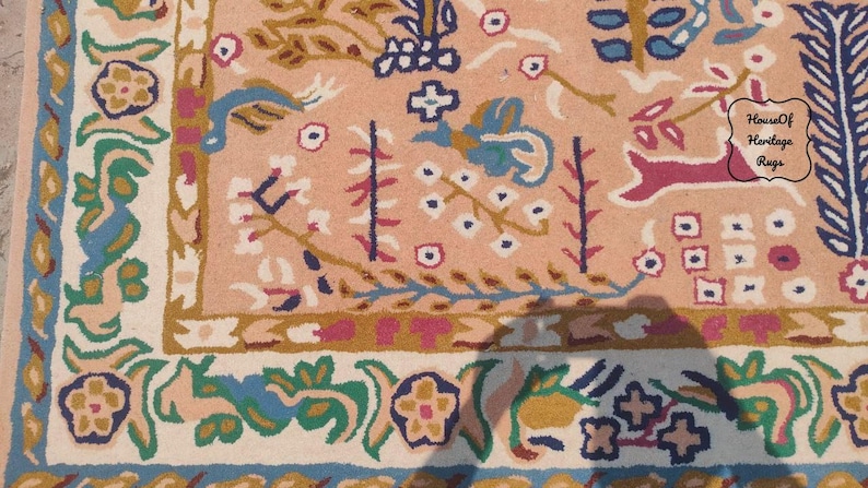 Peut inclure: Gros plan sur une section de tapis avec un fond p&ecirc;che et un motif floral d&eacute;taill&eacute;. Le tapis pr&eacute;sente une bordure avec des accents bleus, verts et jaunes. Le motif central comprend un cerf, des fleurs et d'autres &eacute;l&eacute;ments d&eacute;coratifs. Le texte "House Of Heritage Rugs" est visible.