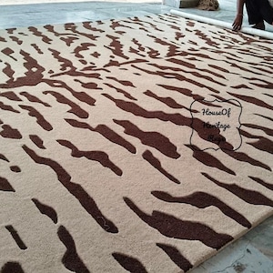May include: A large rectangular area rug featuring a cream-colored base with a dark brown abstract tiger stripe pattern. The rug has a textured pile surface and includes a black outlined label with the text House Of Heritage Rugs.