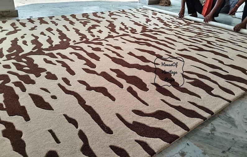 May include: A large rectangular area rug featuring a cream-colored base with a dark brown abstract tiger stripe pattern. The rug has a textured pile surface and includes a black outlined label with the text House Of Heritage Rugs.