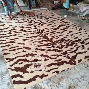 May include: A rectangular area rug featuring a dark brown abstract tiger stripe pattern on a light beige background. The rug includes a black outlined label in the lower center with the text House Of Heritage Rugs printed in a black script font.