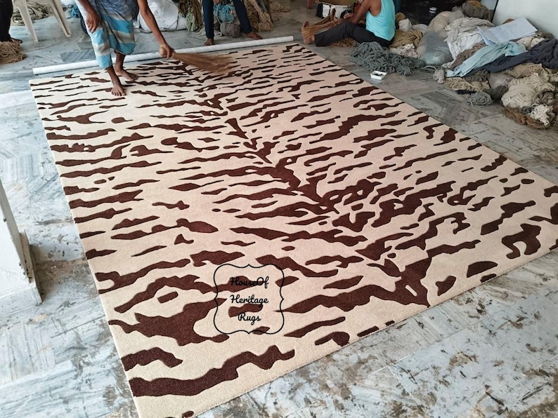 May include: A rectangular area rug featuring a dark brown abstract tiger stripe pattern on a light beige background. The rug includes a black outlined label in the lower center with the text House Of Heritage Rugs printed in a black script font.