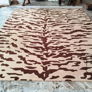 May include: A rectangular area rug featuring a dark brown tiger stripe pattern on a light beige background. The rug has a central spine design with symmetrical markings extending outward. A black logo with the text House Of Heritage Rugs is visible.