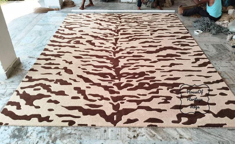 May include: A rectangular area rug featuring a dark brown tiger stripe pattern on a light beige background. The rug has a central spine design with symmetrical markings extending outward. A black logo with the text House Of Heritage Rugs is visible.