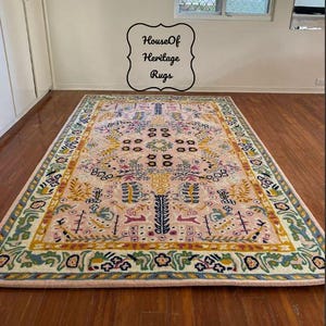 Peut inclure: Un tapis rectangulaire avec un champ central rose et une bordure d&eacute;taill&eacute;e. Le tapis pr&eacute;sente un motif floral et g&eacute;om&eacute;trique complexe dans des tons de jaune, bleu, vert et rose. Le tapis est pos&eacute; sur un sol en bois.