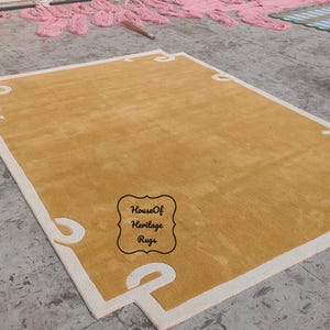 Op de afbeelding: Mosterdgele rechthoekige vloerkleed met een witte rand en decoratieve hoekaccenten. Het vloerkleed ligt op een betonnen ondergrond. Het vloerkleed heeft de tekst "House Of Heritage Rugs".