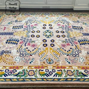 Peut inclure: Un tapis beige avec un motif floral et g&eacute;om&eacute;trique complexe. Le tapis pr&eacute;sente un motif de m&eacute;daillon central avec des accents jaunes, bleus, roses et verts. La bordure pr&eacute;sente un motif r&eacute;p&eacute;t&eacute; d'oiseaux et de fleurs stylis&eacute;s. Le texte "House Of Heritage Rugs" est dans le coin sup&eacute;rieur gauche.